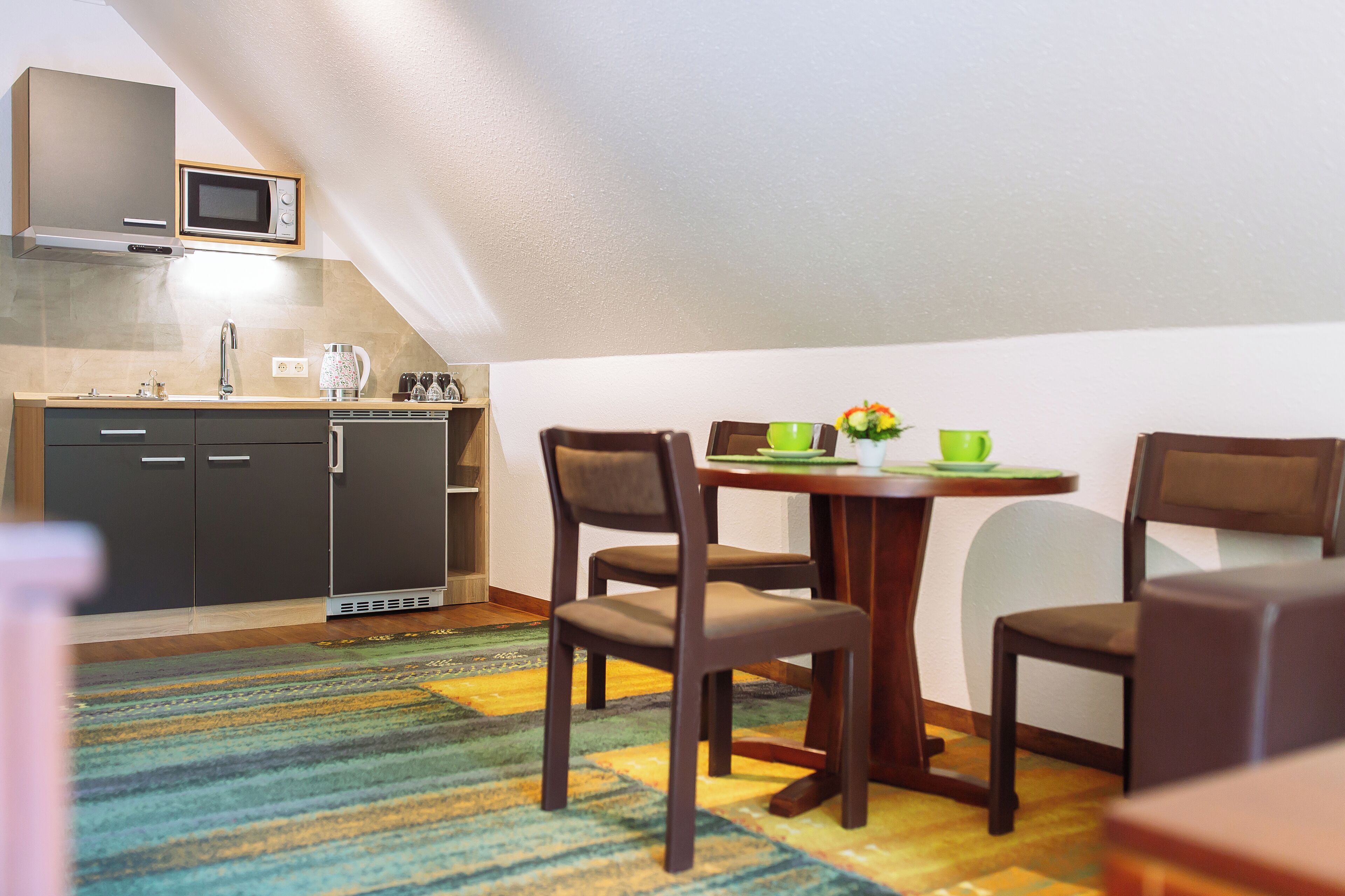 A compact kitchen with a microwave, sink, and cabinets. There is a dining area with a table, chairs, and a colorful rug.