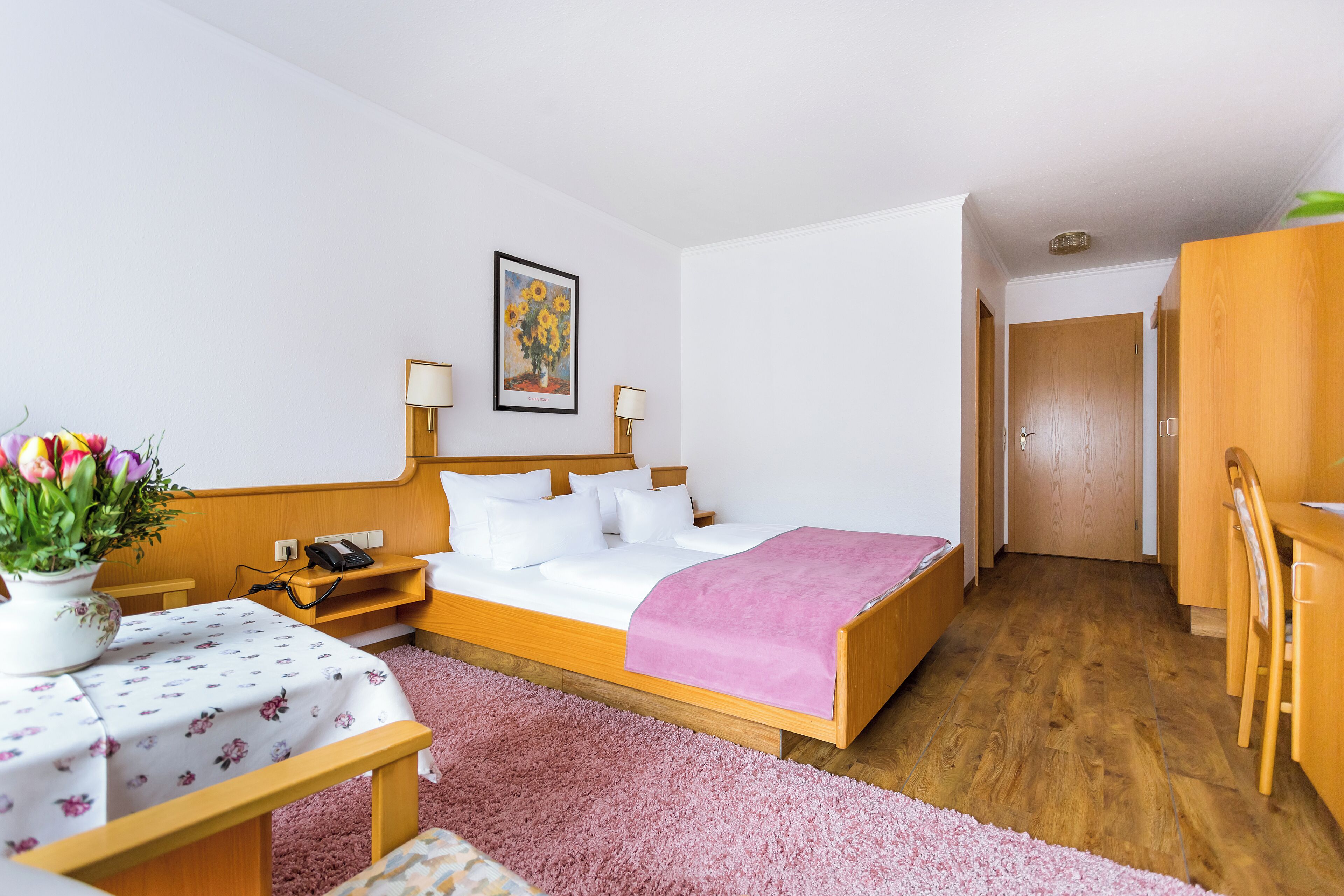 Un dormitorio con cama, mesitas de noche, un escritorio y una silla. Hay una puerta que da a otra habitación.