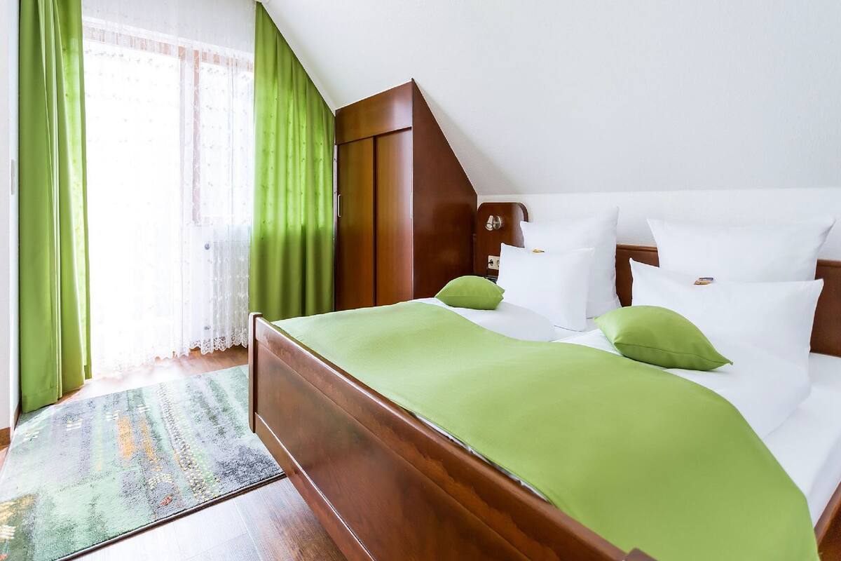 Quarto com cama de madeira, roupa de cama verde, almofadas brancas e vista para um jardim através de uma janela com cortinas verdes.