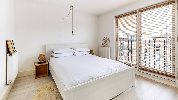 A bedroom with a white bed, wooden nightstands, a hanging light fixture, and a view through large windows with blinds.
