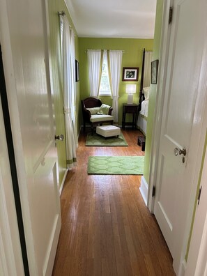 A hallway leading to a room with a green wall, a wooden floor, a chair, a small table, and a window with curtains.