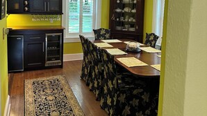 Uma sala de jantar com mesa de madeira, cadeiras e um candelabro. Há também uma cozinha com armários e geladeira.