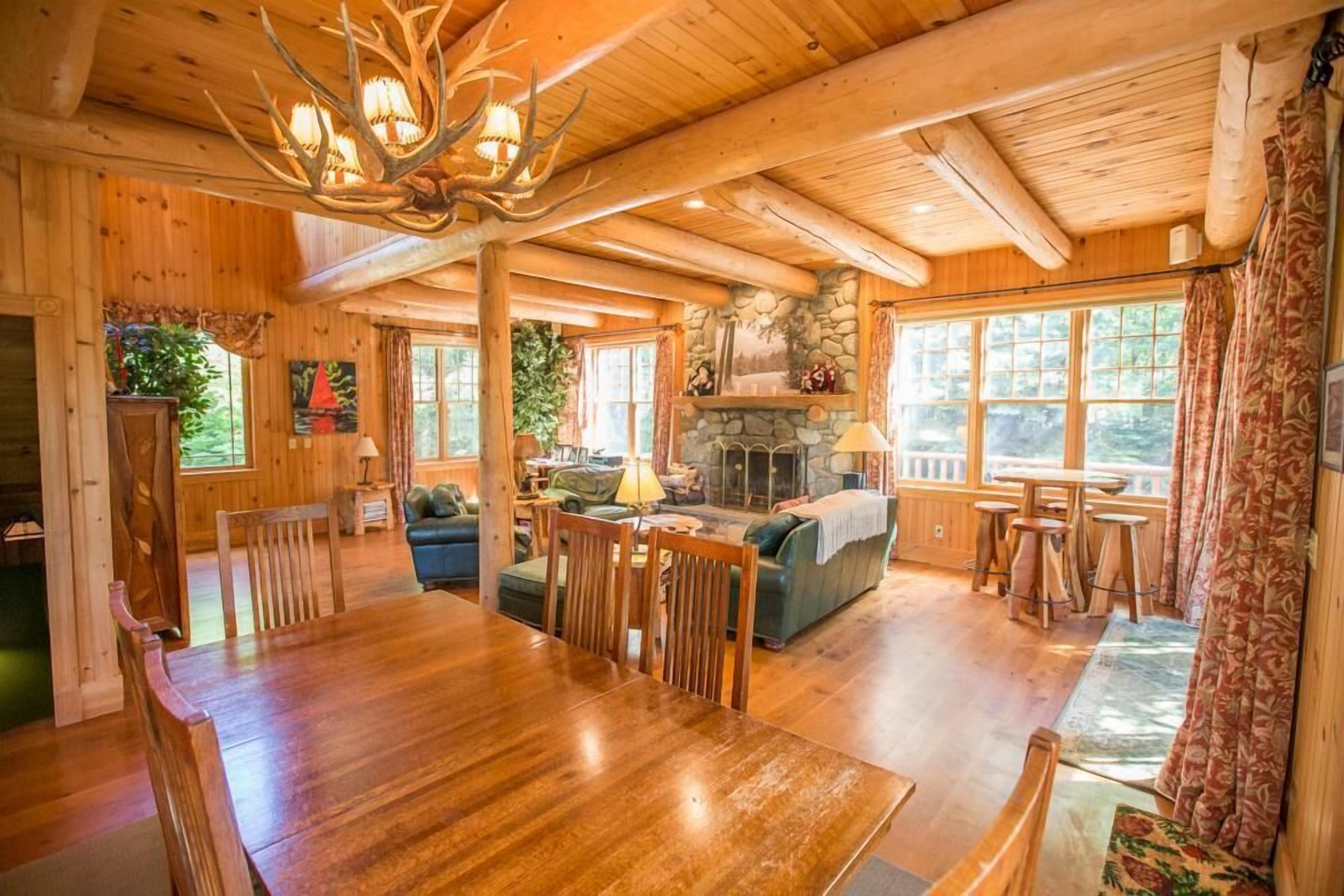 A wooden cabin interior with a dining area, fireplace, and large windows.