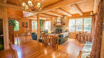 A wooden cabin interior with a dining area, fireplace, and large windows.