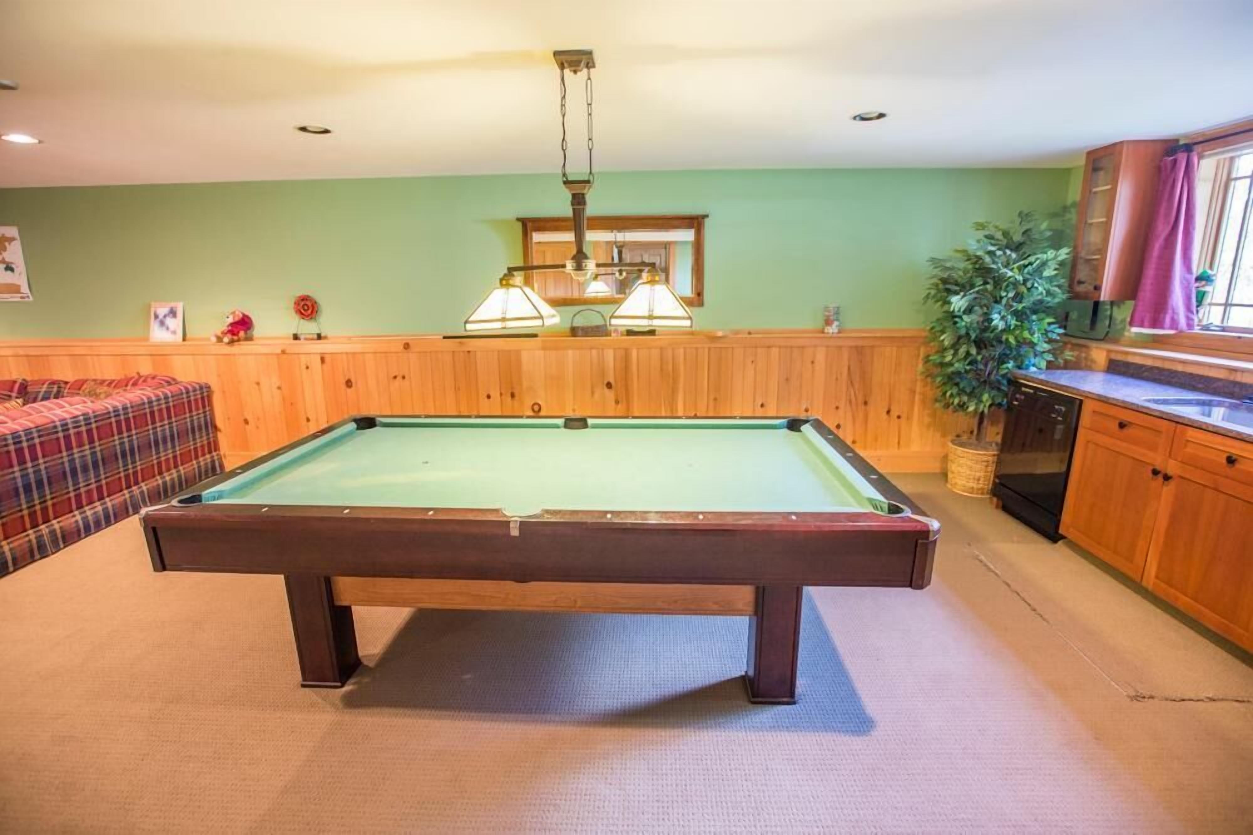 A pool table in a room with wooden paneling, a sofa, and a kitchen area.