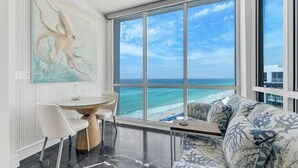 A modern living room with a large window overlooking the ocean, a round glass table with chairs, and a sofa with a floral pattern. - Neptune's Niche 1122 managed by Destin Pointe Vacation Rentals (Destin)