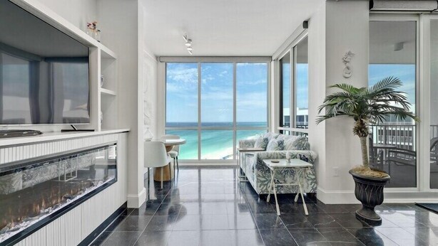 A modern living room with a view of the ocean, a kitchen area, and a balcony with a potted plant. - Neptune's Niche 1122 managed by Destin Pointe Vacation Rentals (Destin)