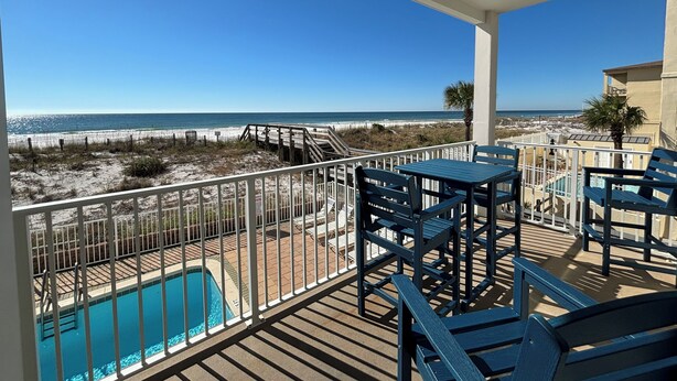 A balcony with a view of the beach, a pool, and outdoor furniture.