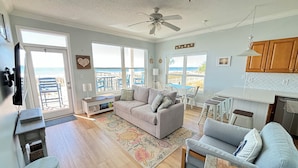 A spacious living area with a large window overlooking a beach, a ceiling fan, and a kitchenette.