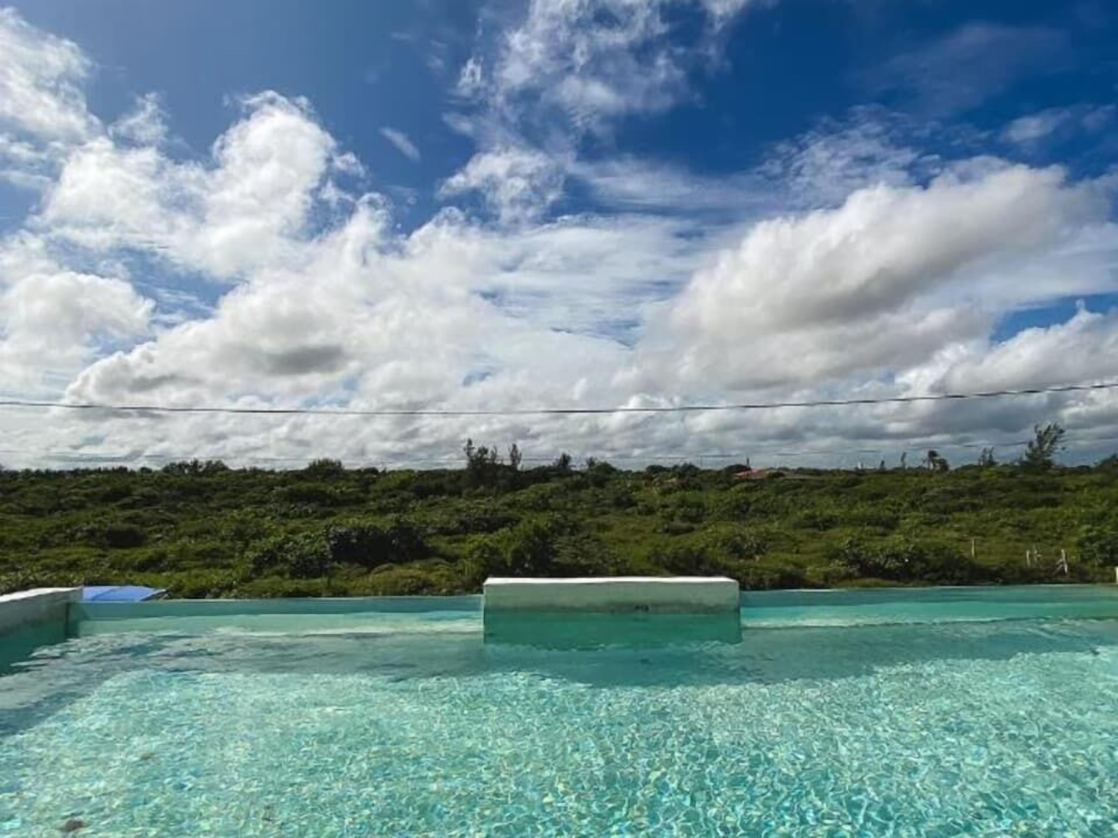 Un cielo azul despejado con nubes, una piscina y un verde cerro en el fondo.