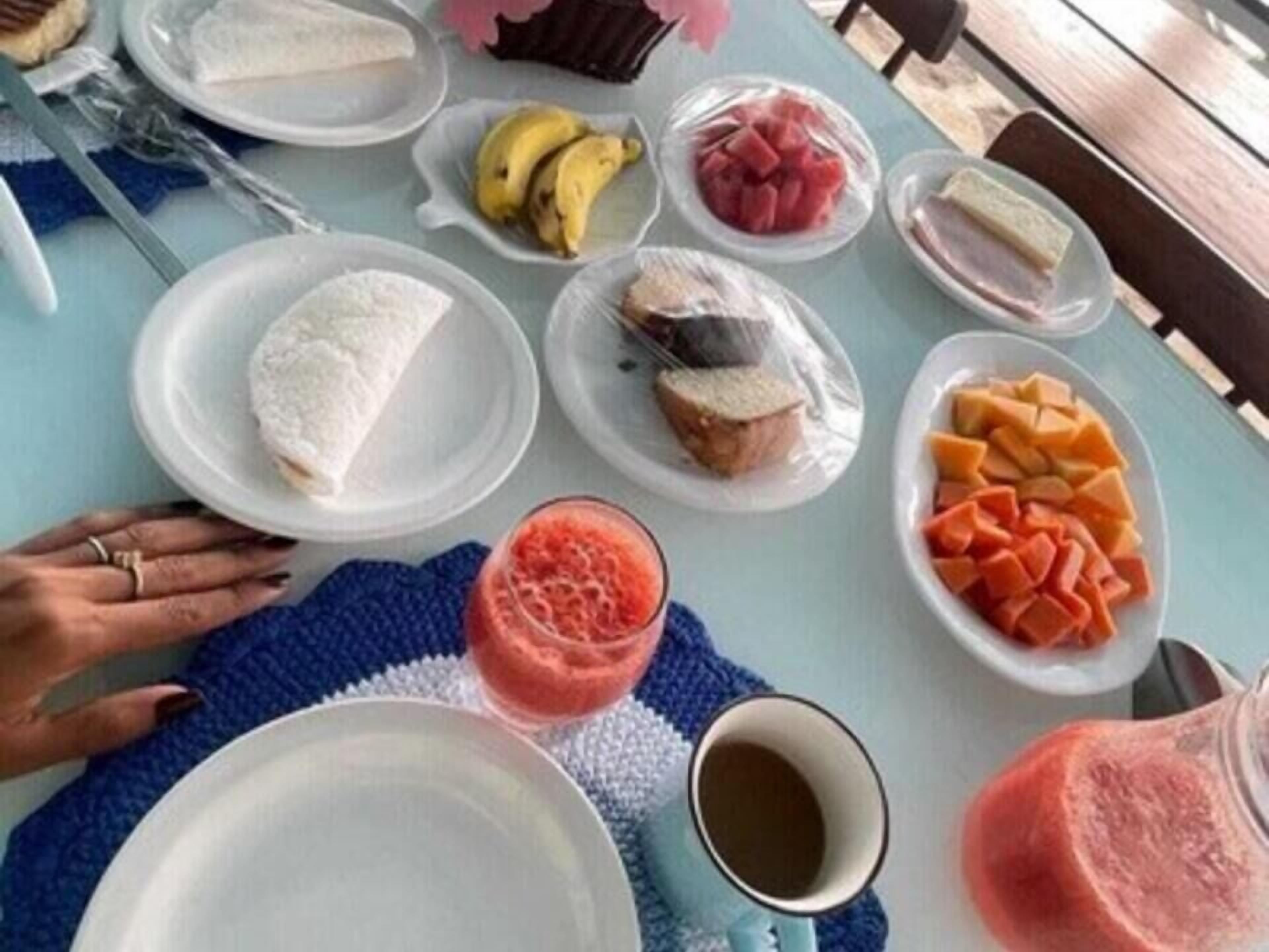 Une table est dressée avec divers aliments et boissons, notamment une assiette de riz blanc, une assiette de tranches de bananes, une assiette de pastèque, une assiette de fromage, une assiette de melon, une assiette de tranches d’orange et un verre de smoothie rose.
