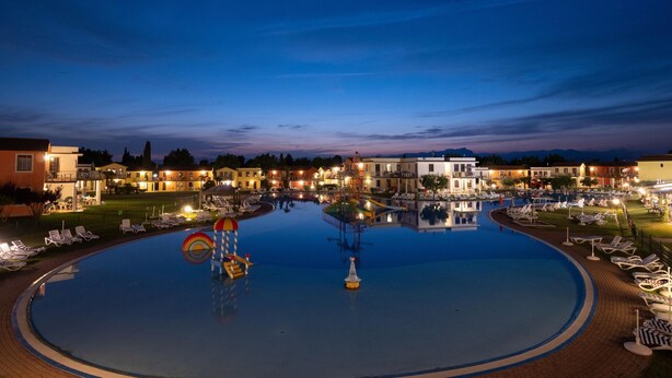 A large outdoor swimming pool with lounge chairs and a water slide, surrounded by buildings with outdoor lighting.
