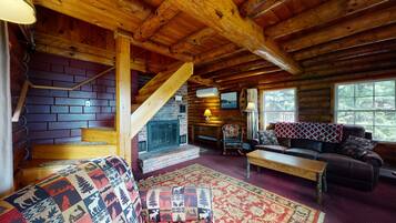 Interior de una cabaña de troncos con chimenea, escalera de madera, sofá de cuero y alfombras estampadas.