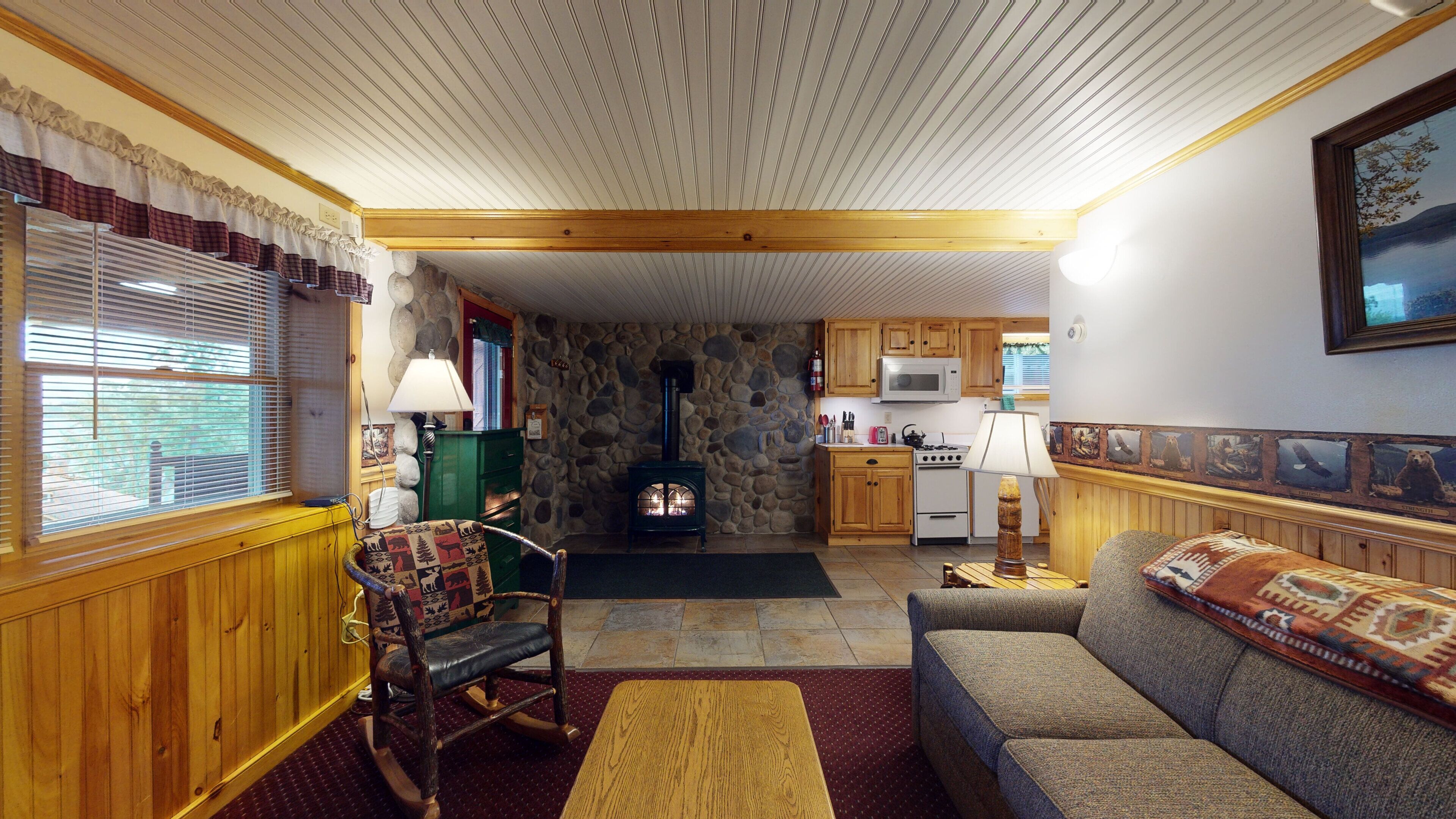 A cozy living room with a stone fireplace, wooden furniture, and a kitchen with wooden cabinets and a microwave.