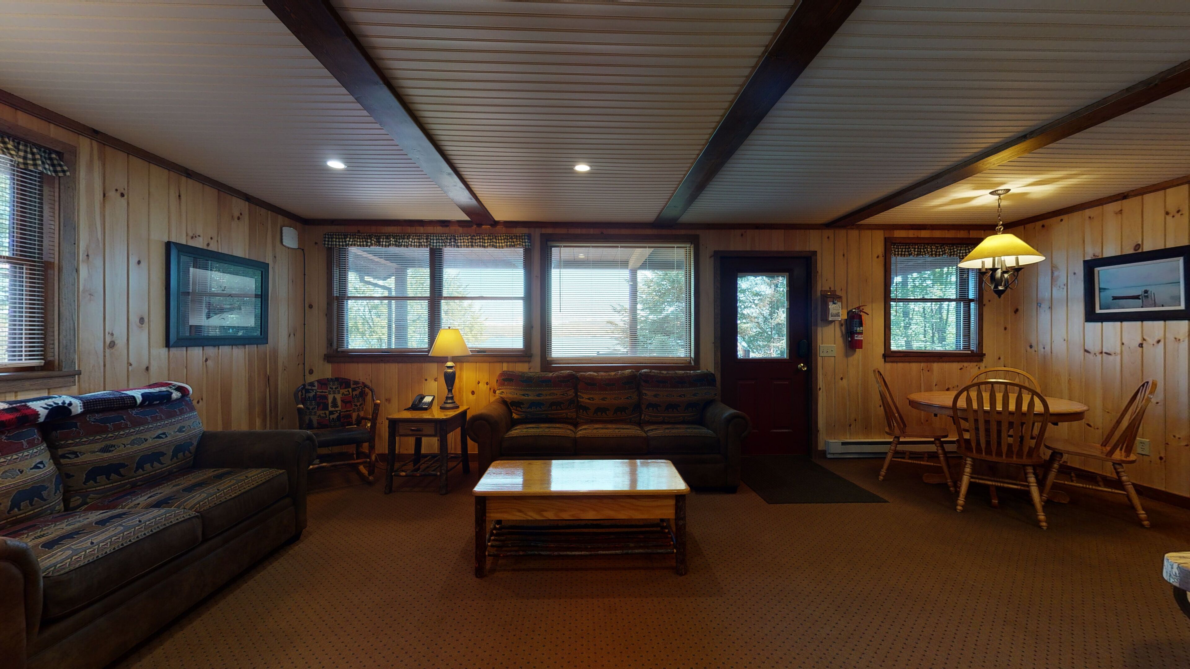 A cozy cabin interior with a wooden floor, a sofa, a coffee table, a dining table with chairs, and large windows with blinds.