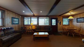 A cozy cabin interior with a wooden floor, a sofa, a coffee table, a dining table with chairs, and large windows with blinds.