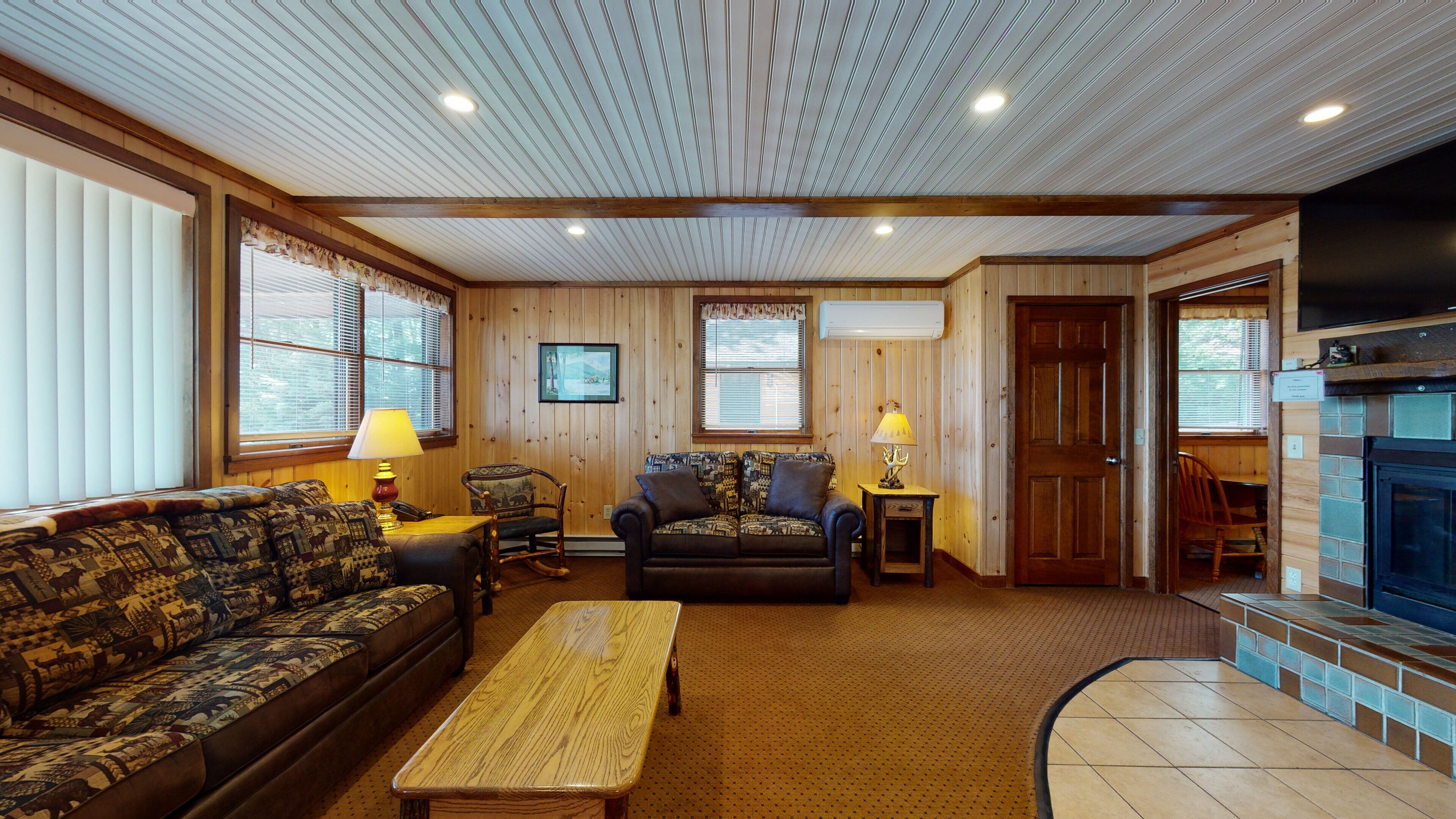 A cozy living room with a sofa, armchair, and coffee table, wooden walls, and a fireplace.