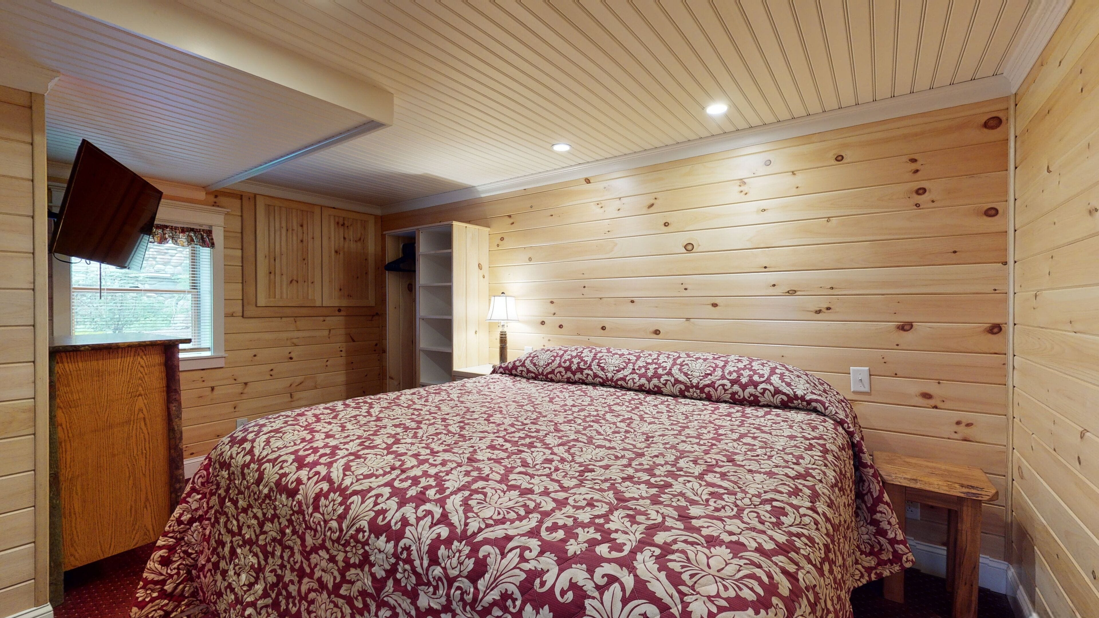 A wooden-paneled bedroom with a bed, a TV mounted on the wall, and a window with blinds.