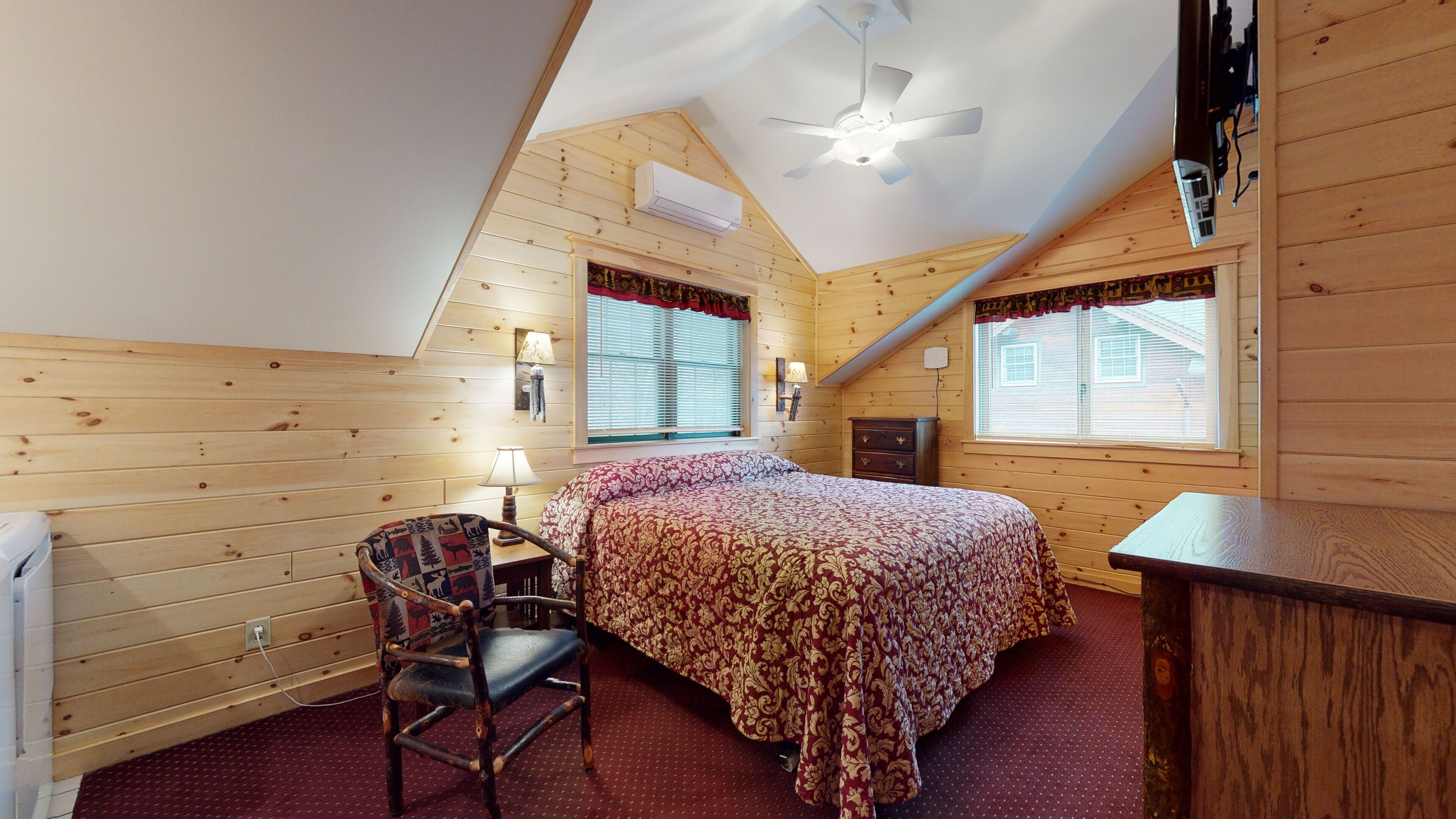 A cozy bedroom with a sloped ceiling, a bed with a patterned bedspread, a chair, a desk, and a window with blinds.
