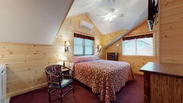 A cozy bedroom with a sloped ceiling, a bed with a patterned bedspread, a chair, a desk, and a window with blinds.