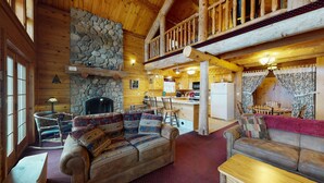 Un acogedor interior de cabaña con chimenea de piedra, muebles de madera, una zona de cocina y un balcón.