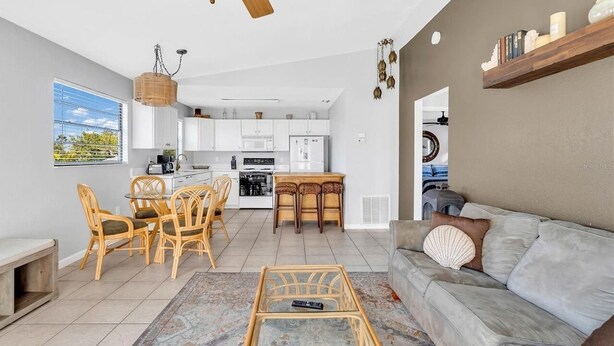 Eine moderne Küche mit weißen Schränken, ein Essbereich mit Korbstühlen und ein graues Sofa mit einem dekorativen Muschelkissen.