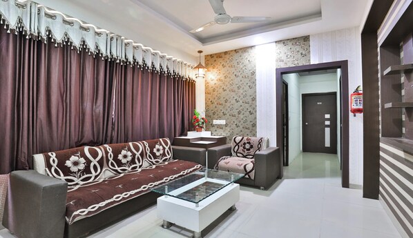 A modern living room with a sofa, a glass coffee table, and a side table. There is a ceiling fan, a wall-mounted lamp, and a door with a fire extinguisher. - Hotel Nandani (Ahmedabad)