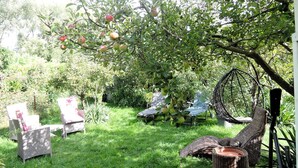 Un jardín con un árbol frutal, sillas de mimbre y una mecedora colgante.