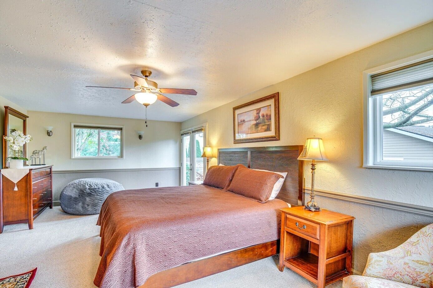 Une chambre à coucher comprenant un lit, une table de chevet, une lampe, un ventilateur de plafond, une commode et une fenêtre.
