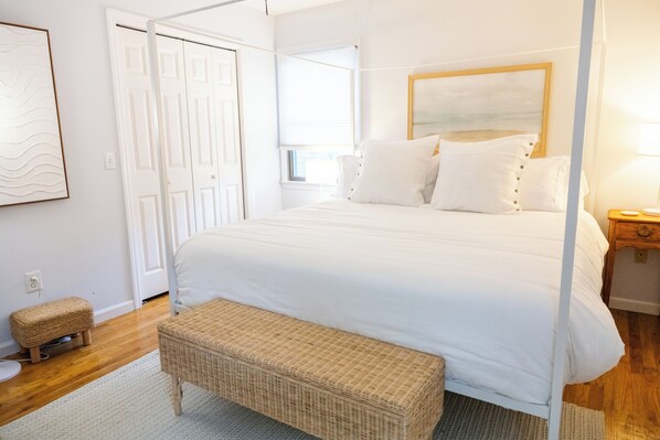 A bedroom with a four-poster bed, a wicker bench, a small stool, and a nightstand with a lamp.