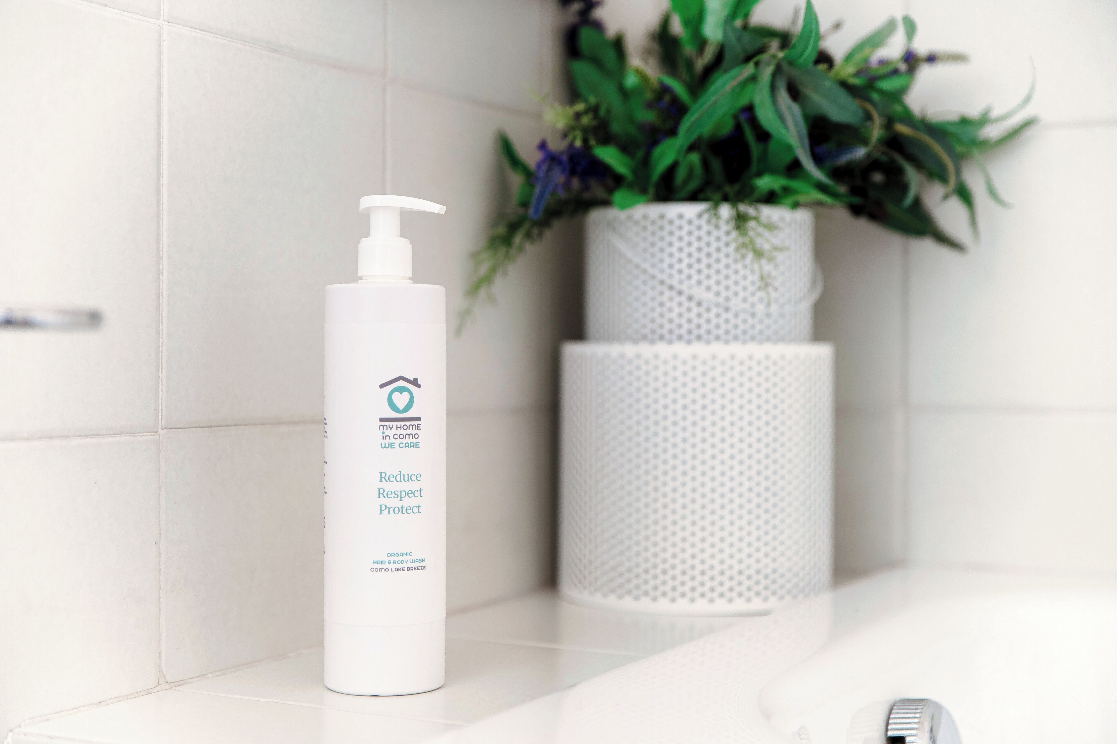 A bathroom with white tiles, a white bottle of organic shampoo, and a white plant in a textured pot.