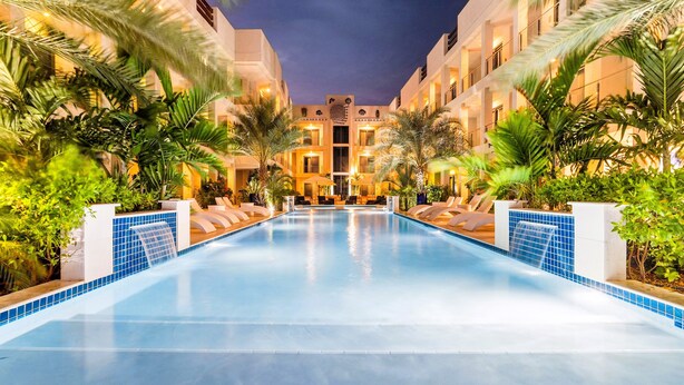 A swimming pool surrounded by palm trees and lounge chairs, with a building in the background. - Bocobay Wariruri Condos (Noord)