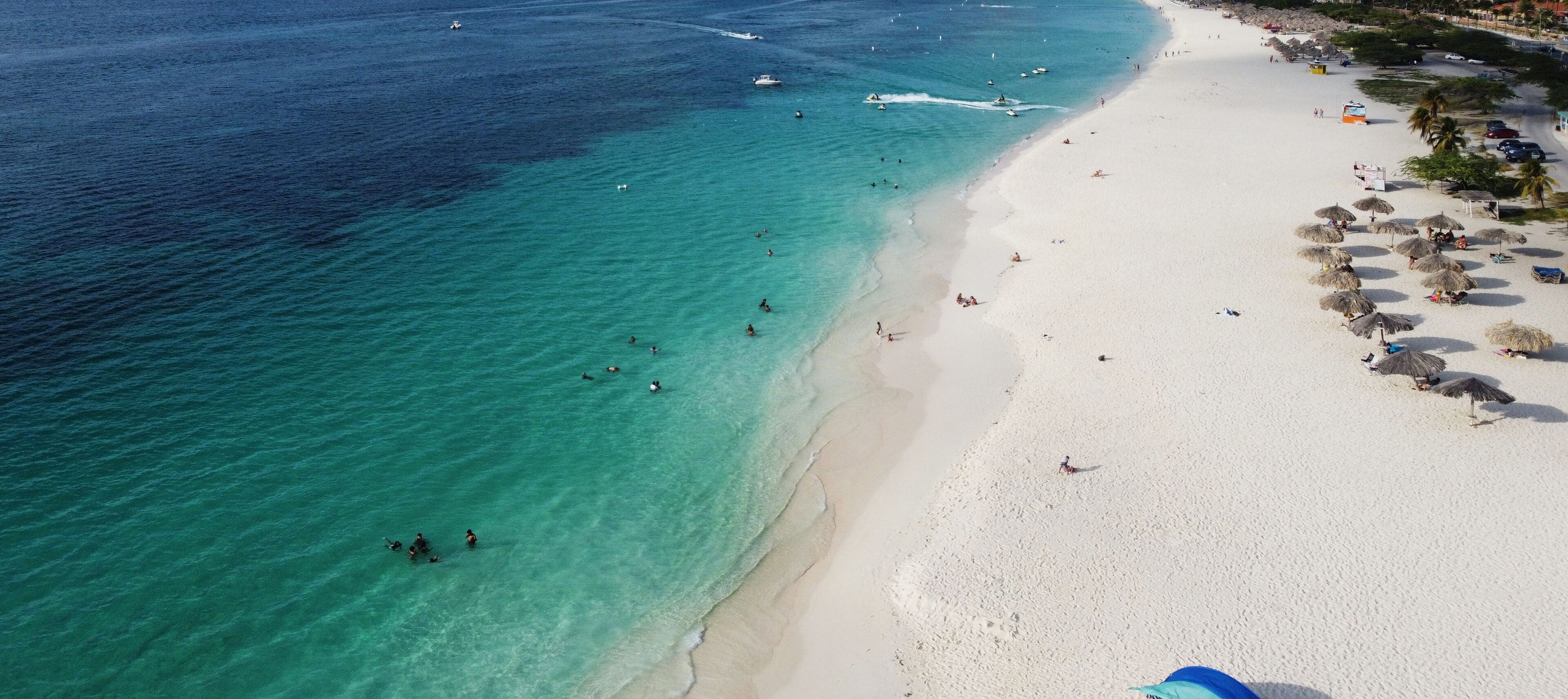Eine Luftaufnahme eines Strandes mit klarem türkisblauem Wasser, weißem Sand und Menschen, die schwimmen und sich sonnen.