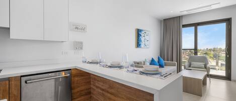 A modern kitchen with a dishwasher, wooden accents, a central island with diningware, a sofa, and a view through large windows.
