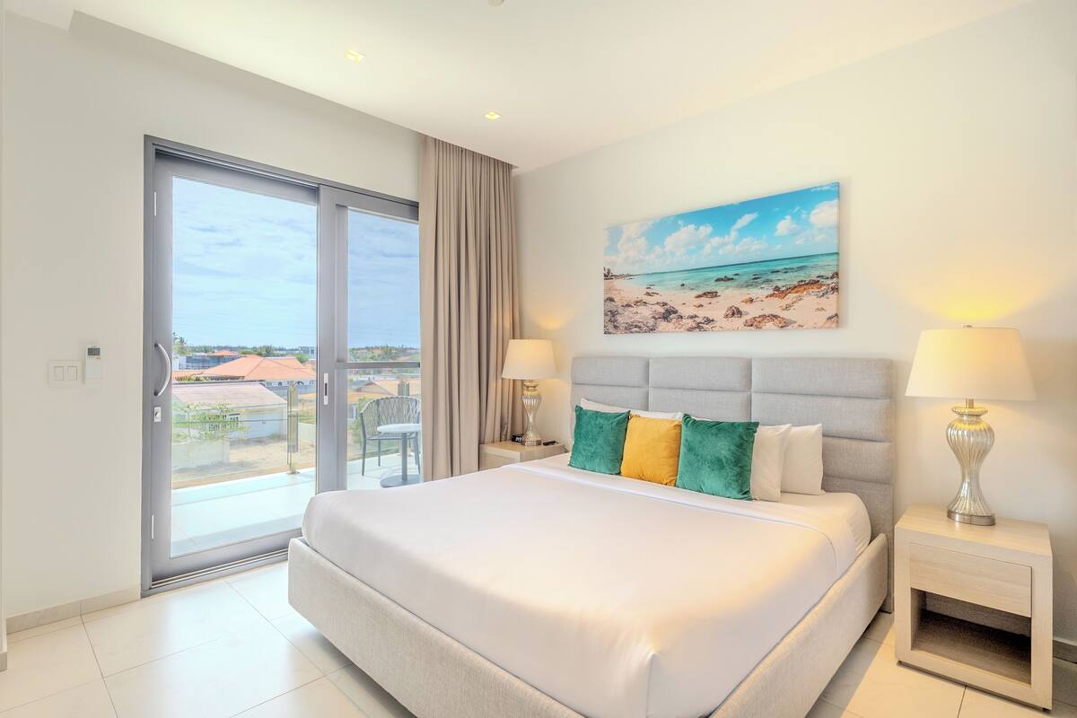 Ein Hotelzimmer mit einem großen Bett, zwei Nachttischlampen, einem Strandbild und einer Schiebetür nach draußen.