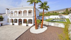 Ein weißes Gebäude mit Balkon, Palmen, Schwimmbecken und einer Kiesfläche.