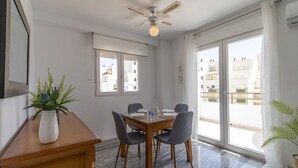 Un coin repas avec une table en bois, quatre chaises, un ventilateur de plafond et une vue sur la ville grâce à de grandes fenêtres.