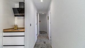 Un couloir étroit, avec un coin cuisine sur la gauche, comprenant une hotte noire, un plan de travail en bois et un petit appareil électroménager. Le sol est carrelé et les murs sont blancs.