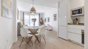 A modern kitchen with a microwave, a dining area with a glass table and white chairs, and a living area with a sofa and a TV.