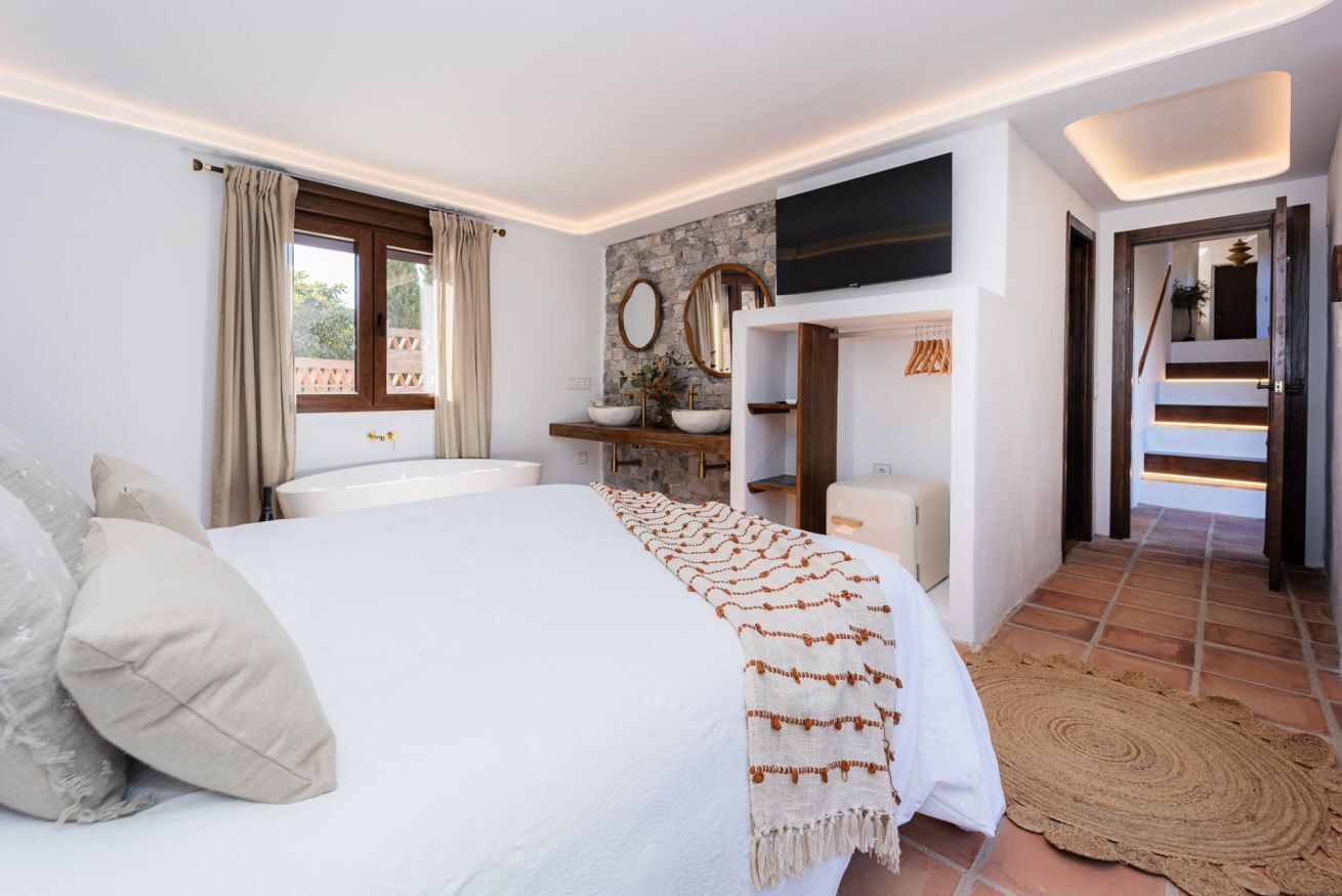 A bedroom with a bed, a bedside table, a TV, and a bathroom with a sink and mirror.