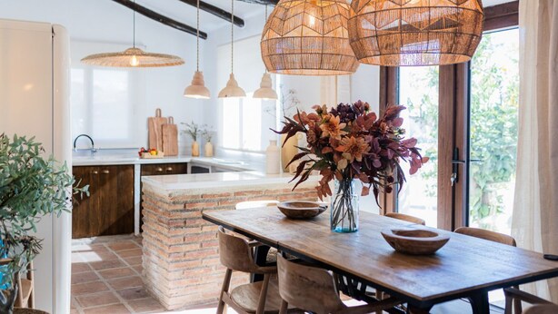Un coin repas avec une table en bois, des suspensions en osier et un vase rempli de fleurs.