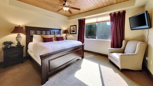 A bedroom with a wooden ceiling, a bed with a headboard, a nightstand, a lamp, a chair, a television, and a window with curtains. - Two levels | Gas fireplace | Walk to ski slopes (Brian Head)