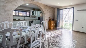 Un comedor con una mesa dispuesta para cuatro personas, sillas blancas, un tapete estampado y una cocina visible a través de un arco.