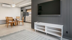 Ein modernes Wohnzimmer mit einem Wand-Fernseher, einem Essbereich mit Holzsesseln und einem Tisch sowie einer Küchenzeile mit Kühlschrank und Herd.