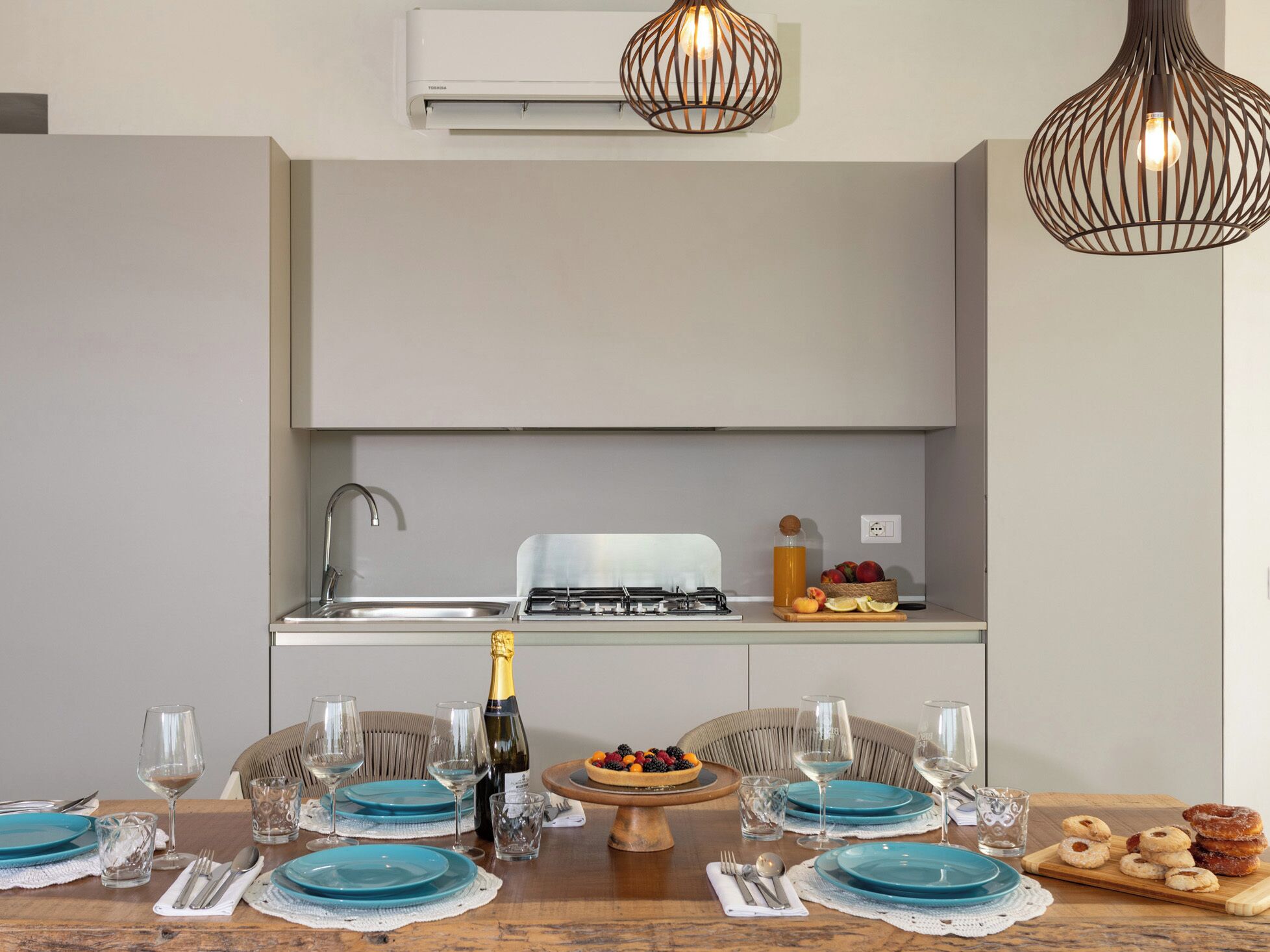 A modern kitchen with a built-in stove, sink, and white cabinets. There is a dining table set with blue plates, glasses, and a bottle of champagne.