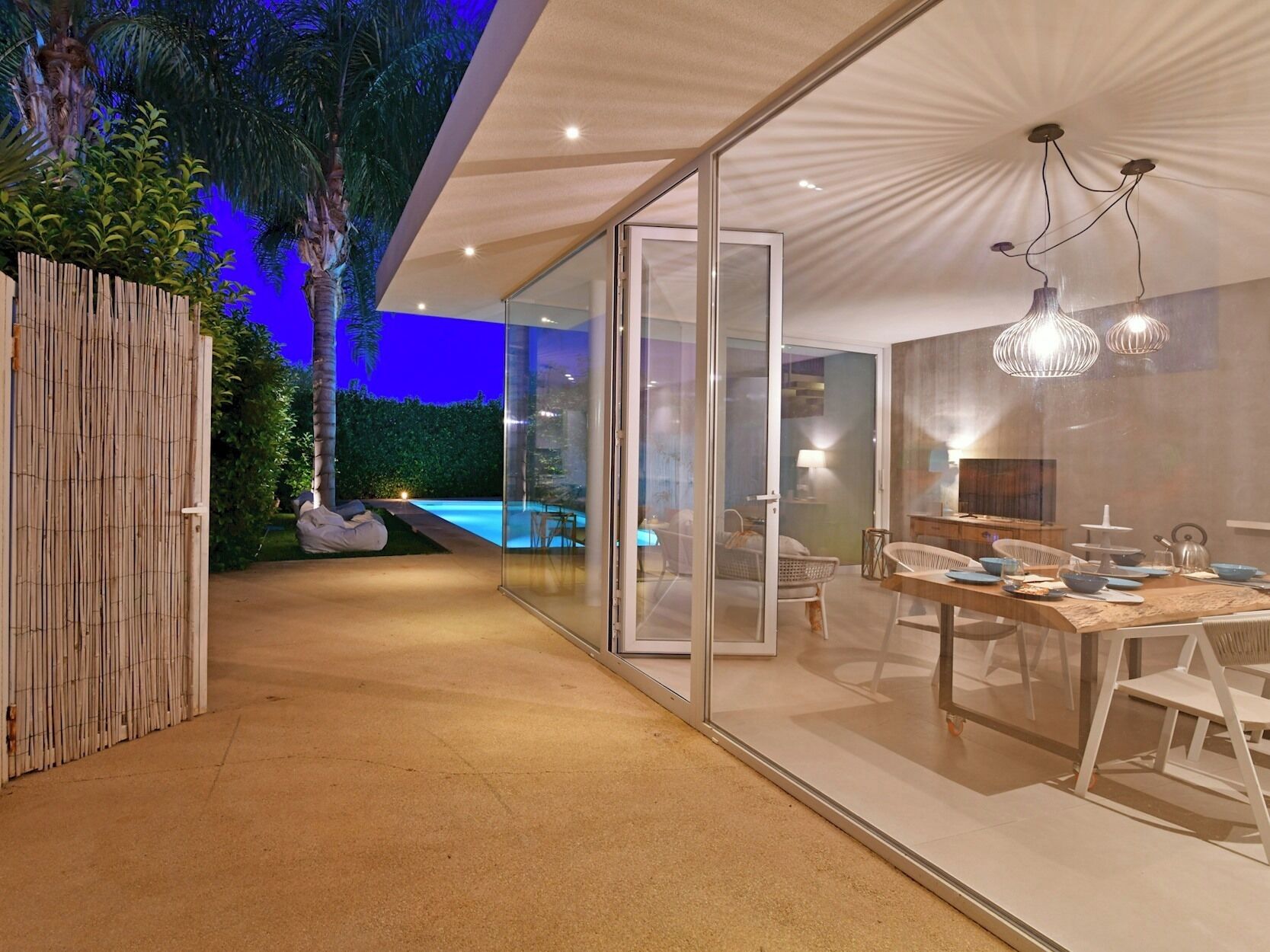 Un interior moderno con un puertas de cristal que da a una zona de piscina, una zona de comedor con una mesa dispuesta para cuatro personas, y un techo con una lámpara de diseño único.