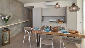 A modern dining area with a wooden table set for four, white chairs, and pendant lighting. - Villa il Melograno 4+2, Emma Villas (Ragusa)