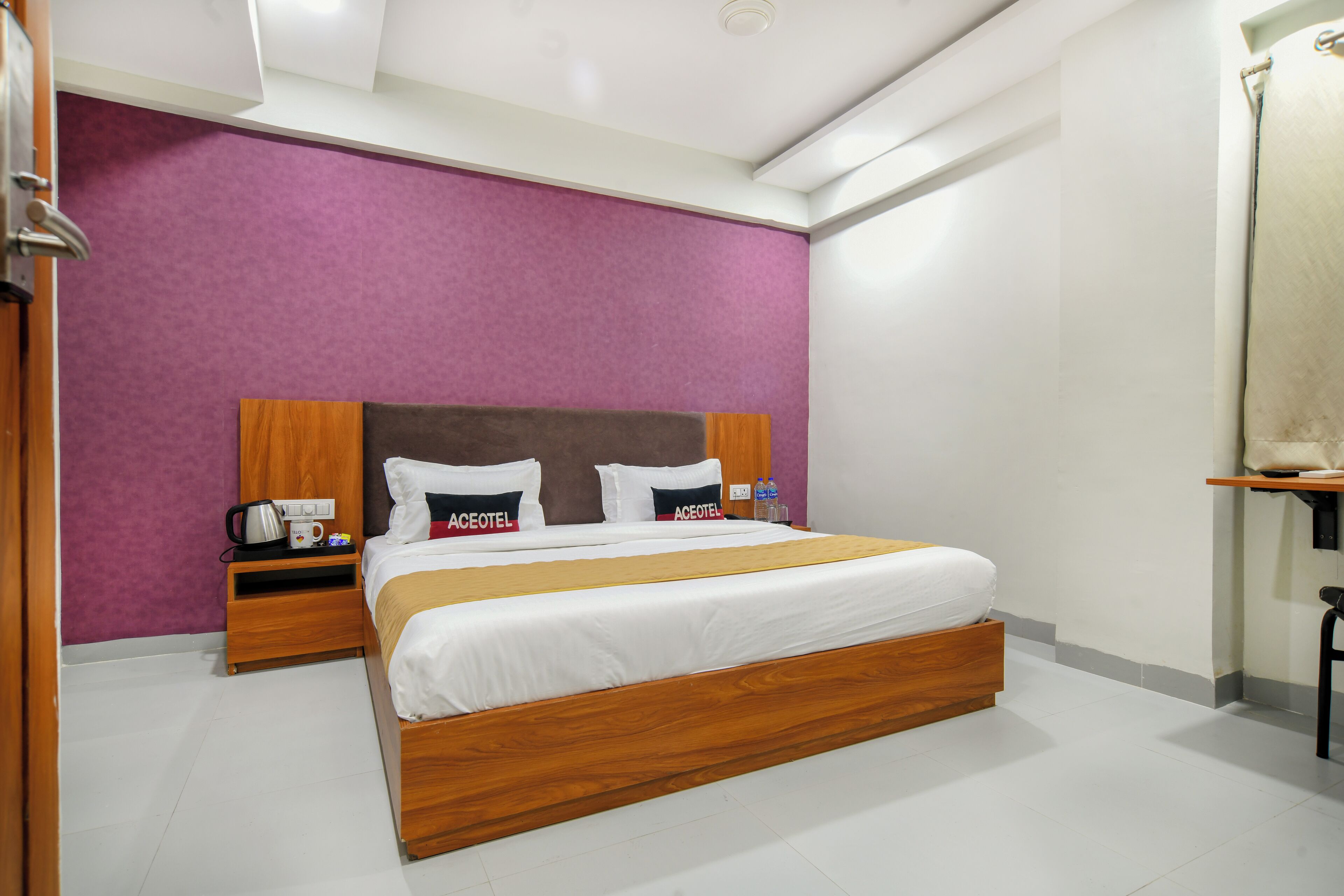 A hotel room with a wooden bed, a purple accent wall, a nightstand with a kettle, and a small table with a chair.
