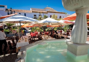 Un espacio de comedor al aire libre con mesas y sombrillas, una piscina y un edificio con techo de tejas al fondo.