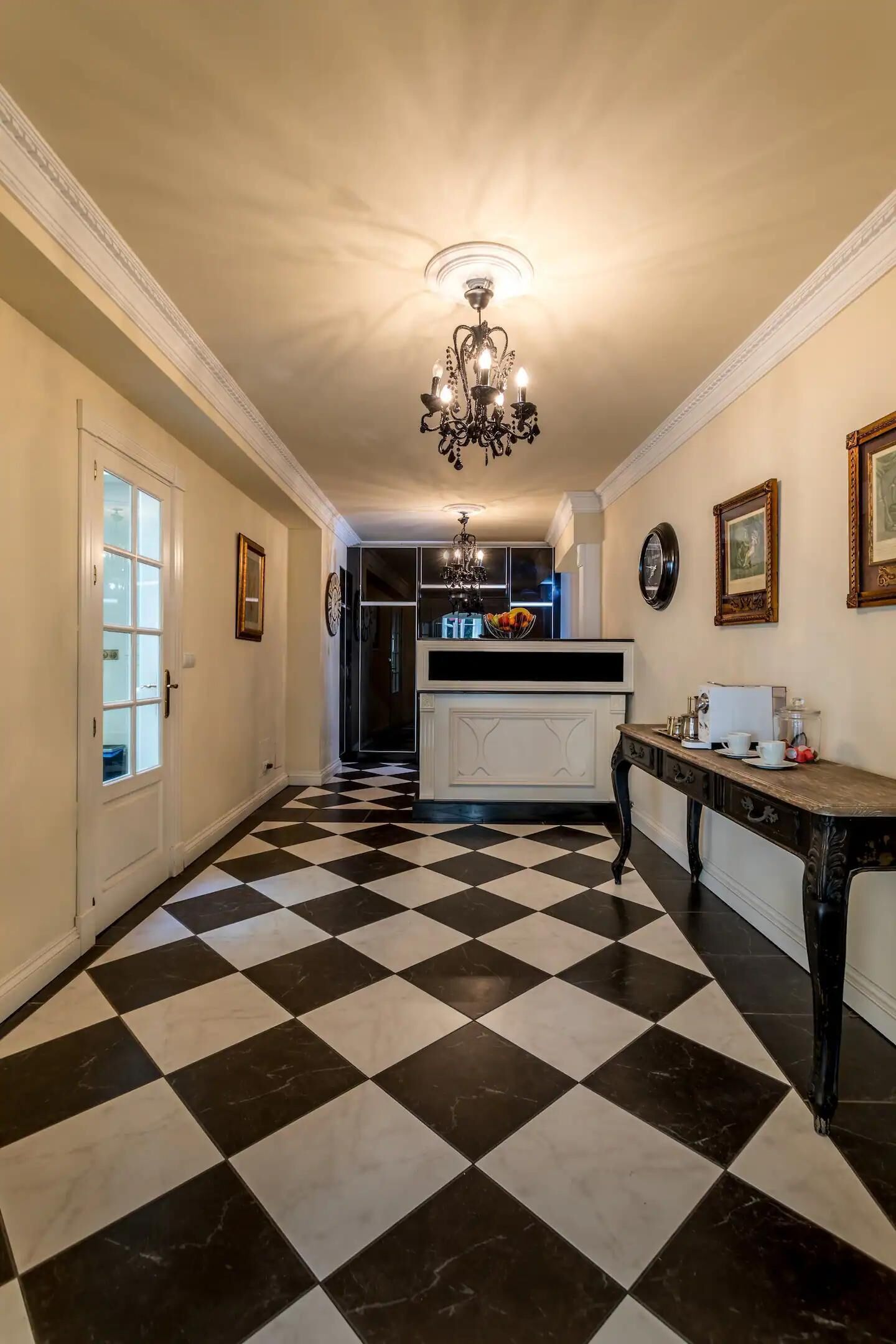 Un pasillo con piso a cuadros blanco y negro, un candelabro, una puerta con paneles de vidrio y una mesa con un jarrón y fruteros.